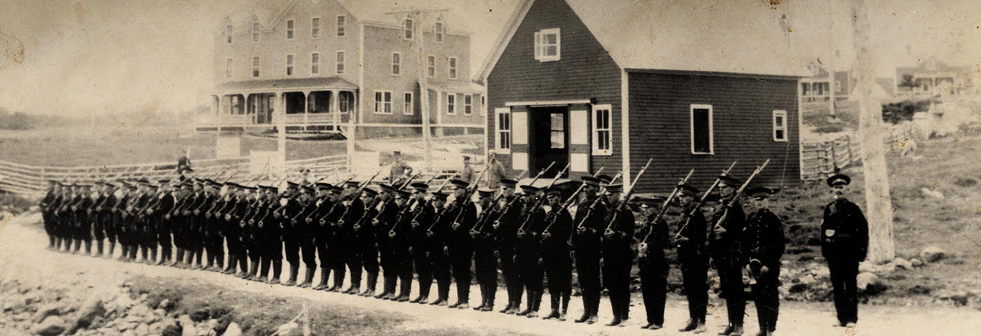 Photographie en noir et blanc de soldats armés debout sur un chemin de terre dans une section rurale de l’île du Cap-Breton. On voit deux bâtiments au loin, dont l’un est la station Marconi.