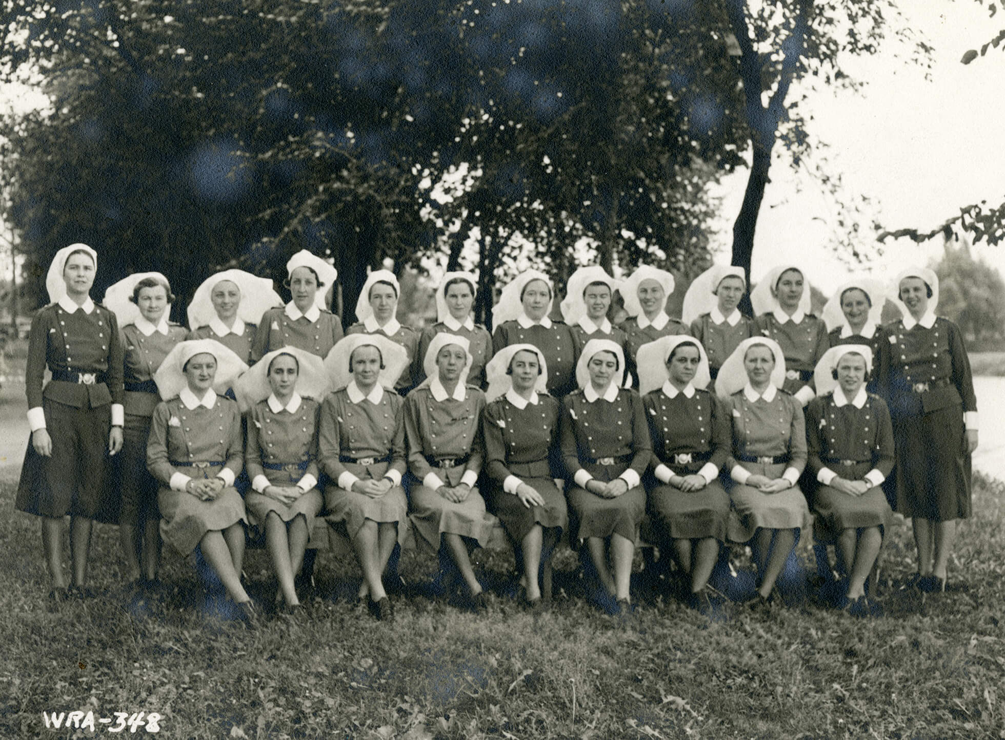 Black and white photograph of a nursing class. Twenty-two women are sitting and standing, wearing nurses' uniforms and white nursing veils. They are outdoors in front of a large tree.