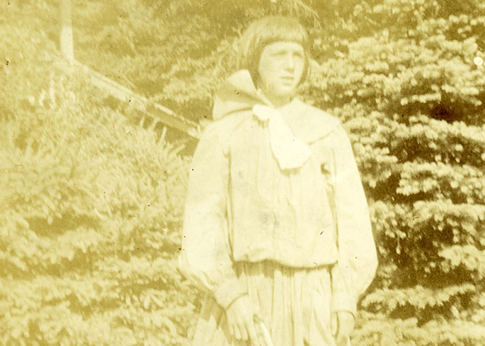 Sepia photograph of a young Helen Kendall standing outdoors holding a gun. She is wearing a dress and sandals. There are trees and a building in the background.