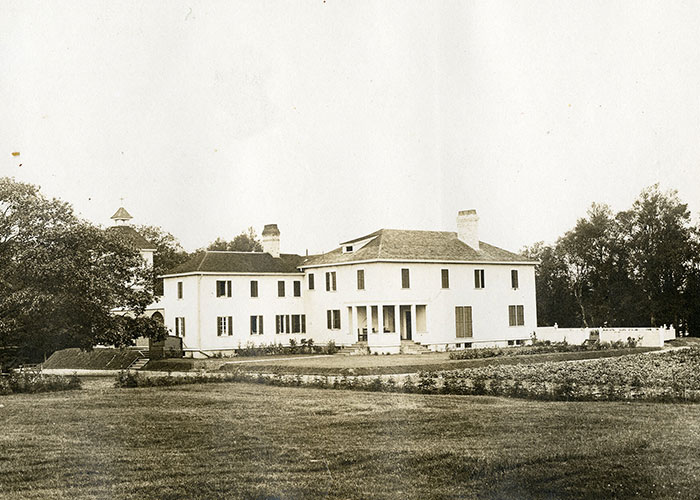 Sepia photograph of a large white house, with a large oak tree on the left hand side of the photograph. The house has two chimneys.