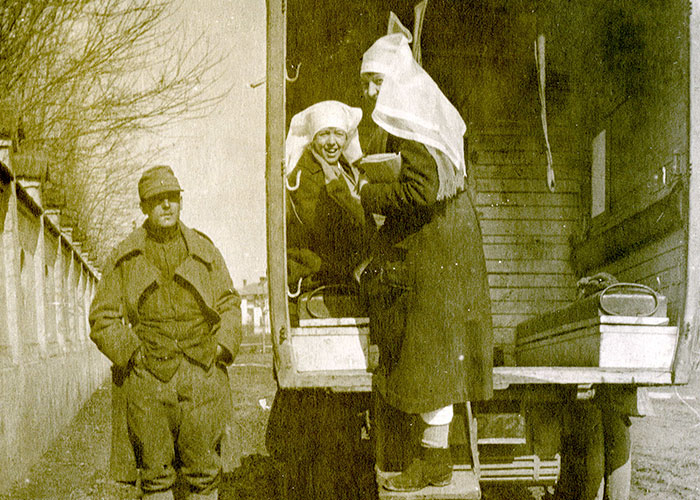 Sepia photograph of a uniformed soldier and two nursing sisters next to a military truck parked on a dirt road. The man is standing at left with his hands in his pockets. One nursing sister is sitting inside the back of the truck and Helen Kendall is standing on the step of the truck, smiling and facing the photographer.