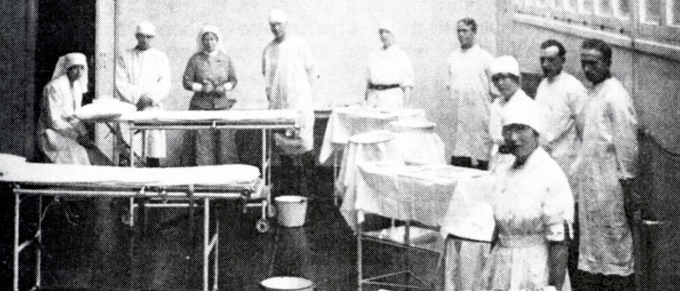 Image is a black and white photograph of personnel in an operating room of a military hospital in England. Several unoccupied patient beds and pieces of surgical equipment are visible. Several men and women are featured in hospital dress. Helen Kendall is pictured at far left. She is seated at the head of a patient bed in preparation to administer an anaesthetic.
