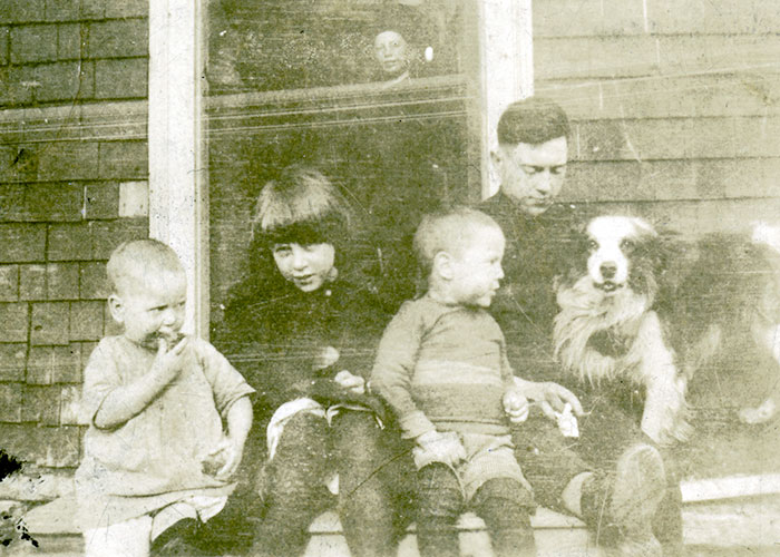 Sepia photograph of an old car. A man is posed at the drivers wheel with a child on his lap. There is a woman and a small child in the back of the automobile. A young boy smiles for the camera while standing outside of the car by its drivers door.