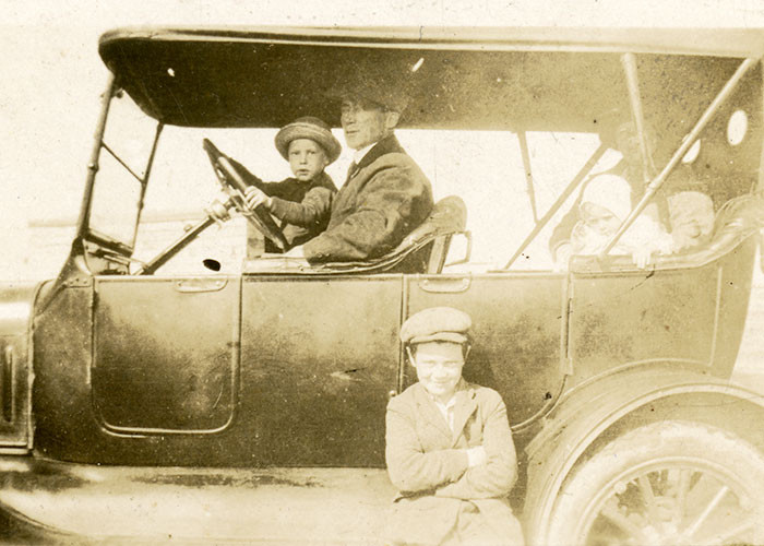 Sepia photograph of an old car. A man is posed at the drivers wheel with a child on his lap. There is a woman and a small child in the back of the automobile. A young boy smiles for the camera while standing outside of the car by its drivers door.
