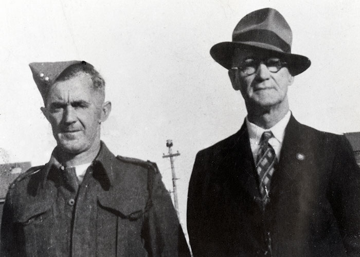 Black and white photograph of two men. The man on the left is wearing a uniform from the Second World War. The man's father is pictured at right wearing a hat and suit and tie.