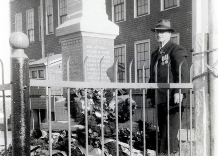 Black and white photograph of a man standing next to a war cenotaph. There are wreaths around the cenotaph and a fence built around the perimeter of cenotaph.